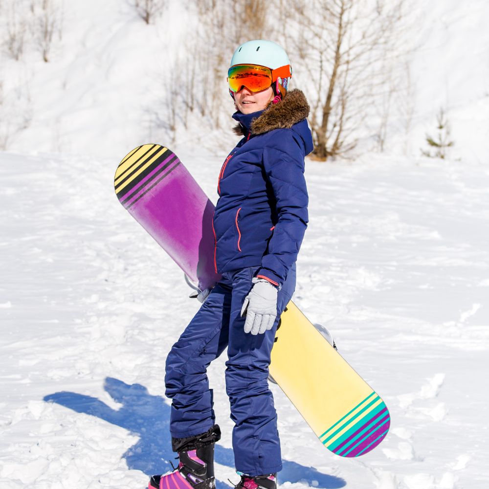 Person in blue snowsuit holding a colorful snowboard in a snowy landscape